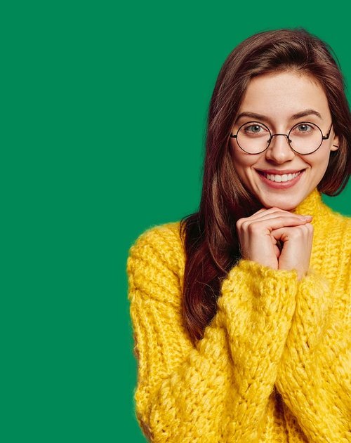 Eine junge Frau mit langen, brünetten Haaren lächelt freundlich in die Kamera. Sie trägt eine auffällige, gelbe Strickjacke und runde Brillengläser. Ihre Hände sind gefaltet und sie zeigt eine enthusiastische oder glückliche Ausdrucksweise. Der Hintergrund ist ein einheitliches, leuchtendes Grün.