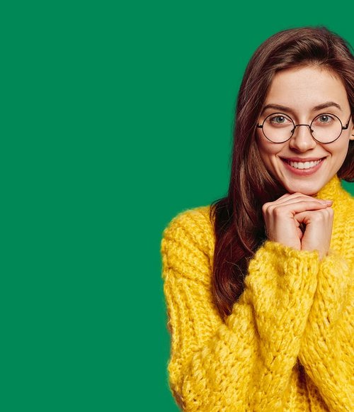 Eine junge Frau mit langen, brünetten Haaren lächelt freundlich in die Kamera. Sie trägt eine auffällige, gelbe Strickjacke und runde Brillengläser. Ihre Hände sind gefaltet und sie zeigt eine enthusiastische oder glückliche Ausdrucksweise. Der Hintergrund ist ein einheitliches, leuchtendes Grün.