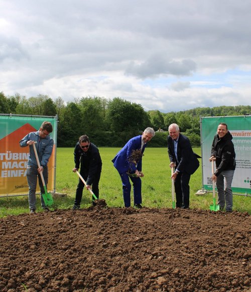 Fünf Personen mit Spaten beim symbolischen Spatenstich auf einem Feld, flankiert von zwei großen Bannern zum Thema Fernwärme. Der Himmel ist bewölkt, im Hintergrund eine grüne Baumlandschaft.