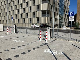 Ein Parkplatz mit gestreifter Pflasterung, auf dem weiße Markierungen für Parkplätze sichtbar sind. Im Hintergrund befindet sich ein modernes Gebäude mit einer Fassade aus senkrechten, perforierten Paneelen. Es gibt mehrere Parkschilder, darunter eines, das auf Ladevorgänge hinweist. Zwei Poller mit roten und weißen Streifen sperren den Zugang, und im Hintergrund sind mehrere Autos zu sehen.