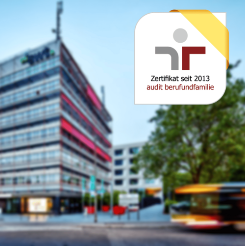 Eine moderne, mehrstöckige Bürogebäude mit einer Glasfassade, die teilweise rot akzentuiert ist. Im Vordergrund stehen einige Bäume und Pflanzen, während sich auf der Straße ein Bus bewegt. In der oberen rechten Ecke ist ein abgerundetes Logo mit einem gelben Band, das die Worte "Zertifikat seit 2013" und "audit berufundfamilie" enthält. Der Himmel ist klar und blau.
