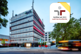 Eine moderne, mehrstöckige Bürogebäude mit einer Glasfassade, die teilweise rot akzentuiert ist. Im Vordergrund stehen einige Bäume und Pflanzen, während sich auf der Straße ein Bus bewegt. In der oberen rechten Ecke ist ein abgerundetes Logo mit einem gelben Band, das die Worte "Zertifikat seit 2013" und "audit berufundfamilie" enthält. Der Himmel ist klar und blau.
