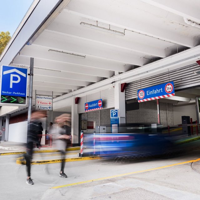 Ein Parkhaus mit einem blauen Parkplatz-Schild "Neckar-Parkhaus". Auf der linken Seite sind ein paar parkende Autos und auf der rechten Seite ist der Eingang zum Parkhaus mit Schildern für "Kasse" und "Eingang". Über der Einfahrt hängt ein weiteres Schild mit der Aufschrift "Einfahrt" und einer Höhenbeschränkung von 1,9 m. Zwei Personen sind in Bewegung, während sie das Parkhaus betreten.