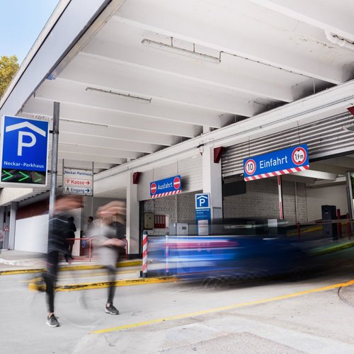 Ein Parkhaus mit einem blauen Parkplatz-Schild "Neckar-Parkhaus". Auf der linken Seite sind ein paar parkende Autos und auf der rechten Seite ist der Eingang zum Parkhaus mit Schildern für "Kasse" und "Eingang". Über der Einfahrt hängt ein weiteres Schild mit der Aufschrift "Einfahrt" und einer Höhenbeschränkung von 1,9 m. Zwei Personen sind in Bewegung, während sie das Parkhaus betreten.