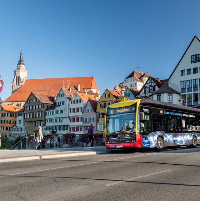 Ein Stadtbus fährt über eine Brücke in einer Stadt mit historischem Fachwerkbau. Auf der linken Seite sind bunte Blumenampeln und dichter grüner Baum. Im Hintergrund sind verschiedene Gebäude mit roten Dächern sichtbar, darunter ein Kirchturm. Es gibt Fußgänger auf der Brücke und Fahrzeuge, darunter ein rotes Auto, auf der Straße. Der Himmel ist klar und blau.