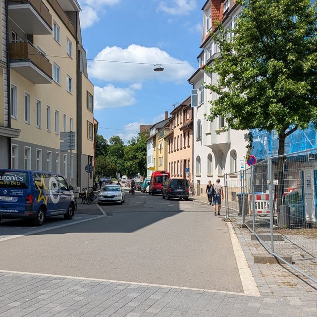 Die Straße ist von mehrstöckigen Gebäuden gesäumt, die überwiegend gelb und beige gestrichen sind. Auf der linken Seite parkt ein blauer Lieferwagen mit dem Aufdruck "euronics Elektro Betz". Diverse Fahrzeuge stehen entlang der Straße, darunter ein roter Bus und mehrere Autos. Einige Personen gehen auf dem Gehweg, während auf der linken Seite ein Radfahrer vorbeifährt. Verkehrszeichen sind sichtbar, darunter ein Schild mit der Geschwindigkeitsbeschränkung von 30 km/h und ein Schild, das Fußgänger und Radfahrer anzeigt. Auf der rechten Seite befindet sich ein Bauzaun und einige Baustellenutensilien sind sichtbar. Der Himmel ist blau mit wenigen Wolken.