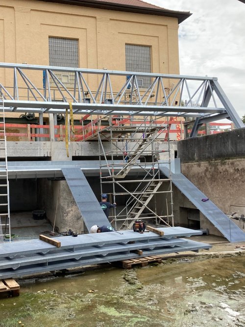 Eine Baustelle mit einem Baugerüst und rampenförmigen Strukturen. Im Vordergrund stehen graue Platten auf Paletten, während ein Arbeiter in einem roten Oberteil auf die Baustelle schaut. Im Hintergrund ist eine Wand mit großen Fenstern und roten Schutzzäunen. Über dem Baugerüst hängen Seile, und es sind Werkzeuge und Materialien sichtbar. Der Boden ist mit Wasser und Pflanzenresten bedeckt.