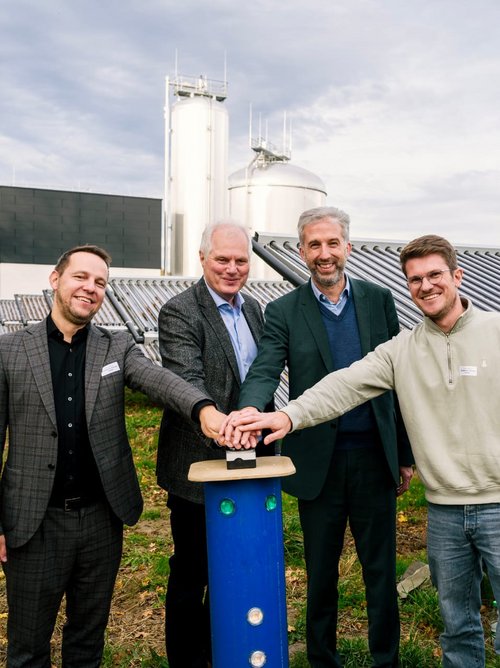 Vier Personen stehen im Freien vor Solarpanelen und einem modernen Gebäude. Sie lächeln und stehen um eine vertikale, blaue Säule herum, auf der ein Knopf montiert ist. Jede Person hat eine Hand auf die Säule gelegt. Im Hintergrund sind weitere Solarpanele sowie zwei große silberne Behälter zu sehen. Die Umgebung ist grün und herbstlich, mit bunten Blättern an den Bäumen.