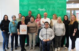 Eine Gruppe von Personen steht und sitzt zusammen in einem Raum, der mit dem Logo der Stadtwerke Tübingen dekoriert ist. Alle Personen lächeln und scheinen glücklich zu sein. Eine Frau hält ein Schild in der Hand, auf dem „TOP LOkalversorger 2023“ steht. Die Gruppe umfasst viele Menschen unterschiedlichen Geschlechts und Alters. Es sind einige Personen in formeller Kleidung und andere in legerer Kleidung.
