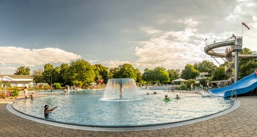 Ein Panoramablick auf ein Freibad mit einem großen Wasserbecken in der Mitte, umgeben von Wasserspielen und einer Rutsche. Menschen genießen den Sommer, schwimmen, spielen im Wasser und entspannen auf Liegen. Eine zentrale Wasserfontäne spritzt Wasser, und im Hintergrund sieht man grüne Bäume und einen blauen Himmel mit einigen Wolken.