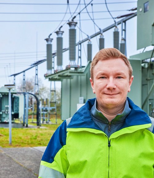 Ein Mann in einer leuchtend gelben und blauen Arbeitsjacke steht in einem Elektrizitätswerk. Im Hintergrund sind Transformatoren und Hochspannungsleitungen zu sehen, und der Himmel ist bewölkt. Die Umgebung wirkt industriell, mit grauem Asphaltboden und einigen Bäumen in der Ferne.