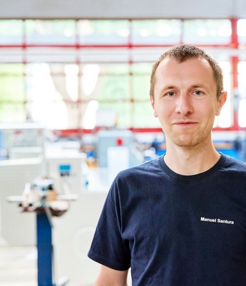 Ein Mann mit kurz geschnittenen Haaren trägt ein schwarzes T-Shirt in einer Werkstatt mit Maschinen im Hintergrund