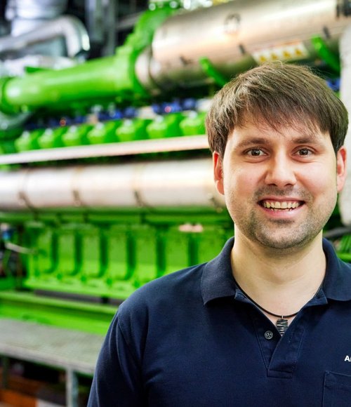 Ein lächelnder Mann steht vor einer großen, grünen Maschinenanlage in einem industriellen Umfeld.