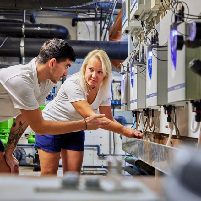 Ein junger Mann und eine junge Frau stehen in einem technischen Raum und betrachten Geräte, die an einer Wand montiert sind. Der Mann ist in einem weißen T-Shirt und kurzer Sporthose gekleidet, während die Frau ebenfalls ein weißes T-Shirt trägt, kombiniert mit einer kurzen Hose. Sie scheinen sich über die Geräte auszutauschen und zeigen auf etwas an der Wand, während im Hintergrund Rohre und grüne Anlagen zu sehen sind.