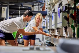 Ein junger Mann und eine junge Frau stehen in einem technischen Raum und betrachten Geräte, die an einer Wand montiert sind. Der Mann ist in einem weißen T-Shirt und kurzer Sporthose gekleidet, während die Frau ebenfalls ein weißes T-Shirt trägt, kombiniert mit einer kurzen Hose. Sie scheinen sich über die Geräte auszutauschen und zeigen auf etwas an der Wand, während im Hintergrund Rohre und grüne Anlagen zu sehen sind.