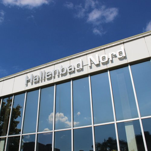Ein modernes Gebäude mit großen Fenstern, auf dem "Hallenbad Nord" in großen, klaren Buchstaben steht. Im Hintergrund ist ein blauer Himmel mit einigen weißen Wolken zu sehen. An der Seite des Gebäudes befinden sich einige grüne Bäume.