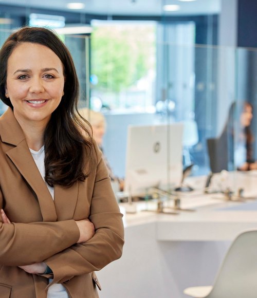 Eine Frau mit langen, braunen Haaren trägt einen beigen Blazer und lächelt, während sie mit verschränkten Armen vor einem modernen Empfangsbereich steht. Im Hintergrund sind weitere Mitarbeitende an Schreibtischen zu sehen, die mit Computern und Glasschutzwänden ausgestattet sind. Der Raum hat große Fenster, durch die Tageslicht einfällt, und ist freundlich und einladend gestaltet.