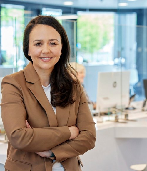 Eine Frau mit langen, braunen Haaren trägt einen beigen Blazer und lächelt, während sie mit verschränkten Armen vor einem modernen Empfangsbereich steht. Im Hintergrund sind weitere Mitarbeitende an Schreibtischen zu sehen, die mit Computern und Glasschutzwänden ausgestattet sind. Der Raum hat große Fenster, durch die Tageslicht einfällt, und ist freundlich und einladend gestaltet.