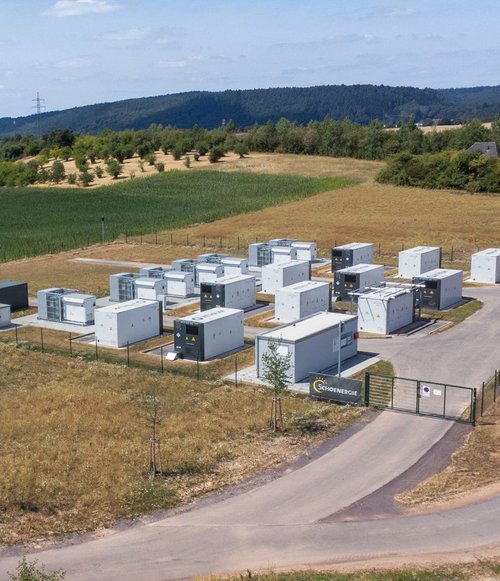 Auf einem Gelände sind mehrere weiße Container und ein paar dunklere Container zu sehen, umgeben von Wiese und Feldern.