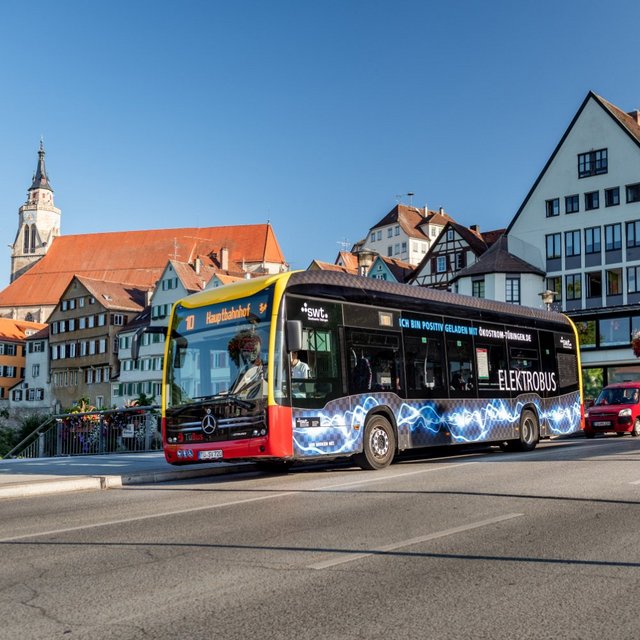Ein Elektrobus fährt über eine Brücke in einer Stadt mit historischer Architektur im Hintergrund. Der Bus hat eine auffällige schwarze Farbgestaltung mit blau leuchtenden Linien und Werbung für elektrische Lösungen. Im Hintergrund sind mehrstöckige Gebäude mit roten und grauen Dächern sowie bunten Blumenkästen zu sehen. Es ist ein sonniger Tag mit klarem blauen Himmel. Auf der Straße fahren mehrere Autos, und einige Personen sind auf Fahrrädern unterwegs.