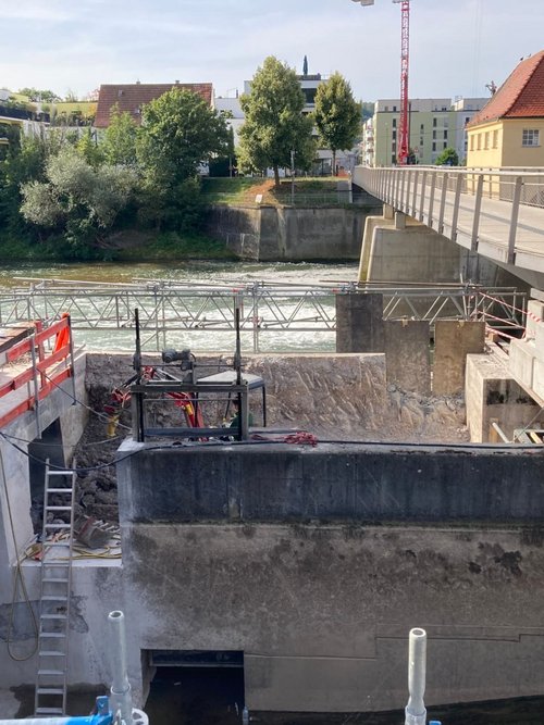 Die Abbildung zeigt eine Baustelle entlang eines Flusses. Auf der linken Seite sind Betonblöcke und ein Gerüst sichtbar, während eine Treppe zu einem tiefer gelegenen Bereich führt. Im Hintergrund fließt der Fluss, und auf der gegenüberliegenden Seite sind mehrere Gebäude, darunter eine gelbe Konstruktion mit einem spitzen Dach, zu erkennen. Über dem Fluss verläuft eine schmale Brücke, die mit einer Geländer versehen ist. In der Umgebung gibt es viele Bäume und Pflanzen, die das Landschaftsbild prägen.
