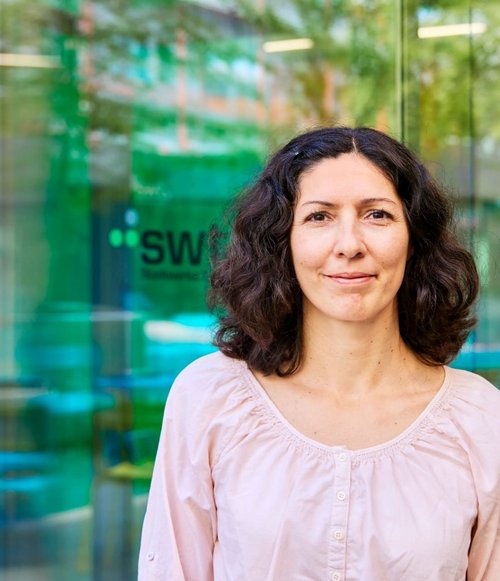 Eine Frau mit lockigem, braunem Haar trägt ein helles Shirt und steht vor einer Glaswand mit reflektierenden Farben.
