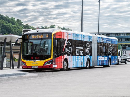 Ein moderner, rot und blau lackierter Linienbus steht an einer Haltestelle. Die Anzeige auf dem Bus zeigt die Route "1" zur "Alxer Straße". Der Bus ist ein E-Bus mit klimafreundlichem Design, das Temperaturen von +4°C und +27°C sowie den Slogan "Wir können nicht bleiben" zeigt. Im Hintergrund sind einige Gebäude und eine andere Fahrzeug auf einer weitläufigen, gepflasterten Fläche zu erkennen. Der Himmel ist bewölkt, und die Umgebung ist von Bäumen umgeben.