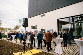 Eine Gruppe von Menschen, die sich an Stehtischen im Freien versammelt hat, umgeben von einem modernen Gebäude. Einige Personen unterhalten sich, während andere Getränke oder Snacks konsumieren. Im Hintergrund sind mehrere Banner und Baupläne sichtbar. Es gibt Tische mit weißen Tischdecken und eine gelbe Plattform am Boden.