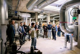 In einem modernen Raum mit großen Metallrohren und Maschinen steht ein Mann, der eine Präsentation hält. Um ihn herum sind mehrere Personen versammelt, die aufmerksam zuhören. Einige haben Kameras oder Smartphones in der Hand, um Fotos zu machen oder Notizen zu machen. Der Raum hat eine industrielle Atmosphäre mit Sichtbeton und gut beleuchteten Bereichen.