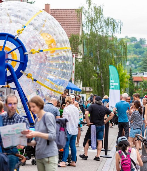 Eine belebte Straße, auf der viele Menschen stehen und sich unterhalten. Im Hintergrund ist ein großes, transparentes Wasserspielgerät zu sehen, das von einer blauen Struktur gehalten wird. In der Nähe gibt es Stände und grüne Banner. Kinder und Erwachsene sind in Gruppen versammelt, einige halten Faltblätter oder Karten in der Hand. Der Himmel ist bewölkt und umgebende Bäume und Gebäude sind im Bild sichtbar.