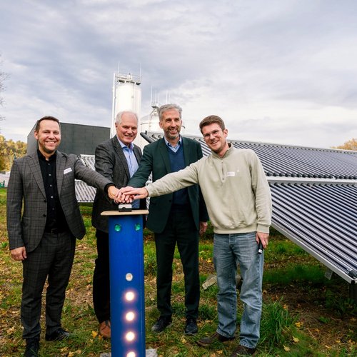 Vier Männer stehen in einem Außenbereich, umgeben von Solarkollektoren. Sie lächeln und haben ihre Hände auf ein vertikales, blaues Gerät gelegt, das beleuchtet ist. Der Hintergrund zeigt eine moderne, graue Gebäude- und Solaranlagenlandschaft, unter einem bewölkten Himmel, während herbstliche Farben in den Bäumen sichtbar sind.