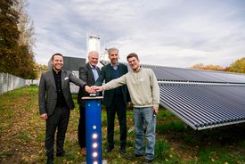 Vier Männer stehen in einem Außenbereich, umgeben von Solarkollektoren. Sie lächeln und haben ihre Hände auf ein vertikales, blaues Gerät gelegt, das beleuchtet ist. Der Hintergrund zeigt eine moderne, graue Gebäude- und Solaranlagenlandschaft, unter einem bewölkten Himmel, während herbstliche Farben in den Bäumen sichtbar sind.