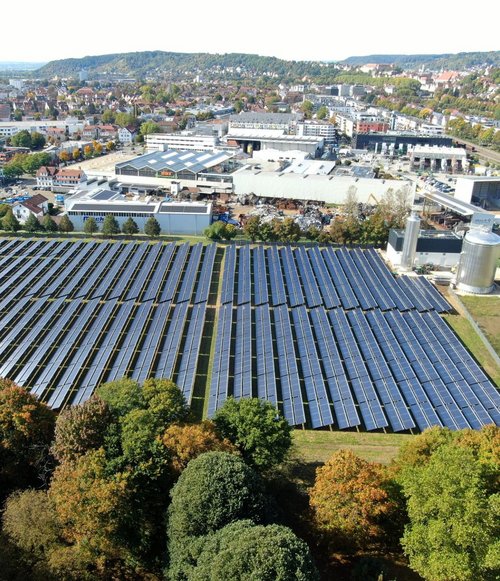 Eine Luftaufnahme zeigt ein großes Solarkraftwerk mit mehreren Reihen von Solarpanelen, die auf einem grünen Gelände installiert sind. Im Hintergrund ist eine Stadtlandschaft mit Wohn- und Geschäftshäusern sowie einer Hauptstraße sichtbar. Die Umgebung besteht aus Bäumen, deren Blätter im Herbst verschiedene Farben annehmen. In der Nähe der Solarpanels sind weitere Gebäude und eine Lagerhalle zu erkennen.