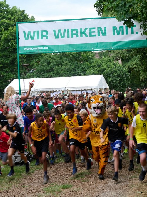 Eine große Gruppe von Kindern und Jugendlichen läuft mit viel Energie beim Start eines Laufes. Einige tragen gelbe und schwarze Sporttrikots, während ein Teilnehmer ein Katzenkostüm, das wie ein Tiger aussieht, trägt. Im Hintergrund sind viele Zuschauer zu sehen, die applaudieren und anfeuern. Eine Person mit blonden Haaren hebt einen roten Trillerpfeifen hoch und gibt den Startschuss. Über der Szene hängt ein Banner mit der Aufschrift "WIR WIRKEN MIT". Es ist eine grüne Landschaft mit Bäumen im Hintergrund sichtbar.