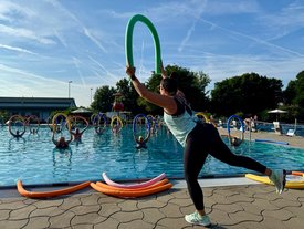 Eine Übungsleiterin zeigt Bewegungen im Freibad, während Teilnehmer sich mit Schwimmreifen im Wasser bewegen.