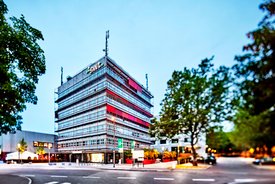 Ein moderner Bürokomplex mit einer Glasfassade in verschiedenen Farbtönen, darunter rot und grau. Das Gebäude hat mehrere Stockwerke und ist umgeben von Bäumen und Strassenlaternen. Im Vordergrund gibt es eine gepflasterte Fläche, auf der sich Fahrradständer befinden. Im Hintergrund sind weitere moderne Gebäude sichtbar, und die Umgebung scheint gut beleuchtet zu sein. Der Himmel ist klar und zeigt eine Dämmerungsstimmung.