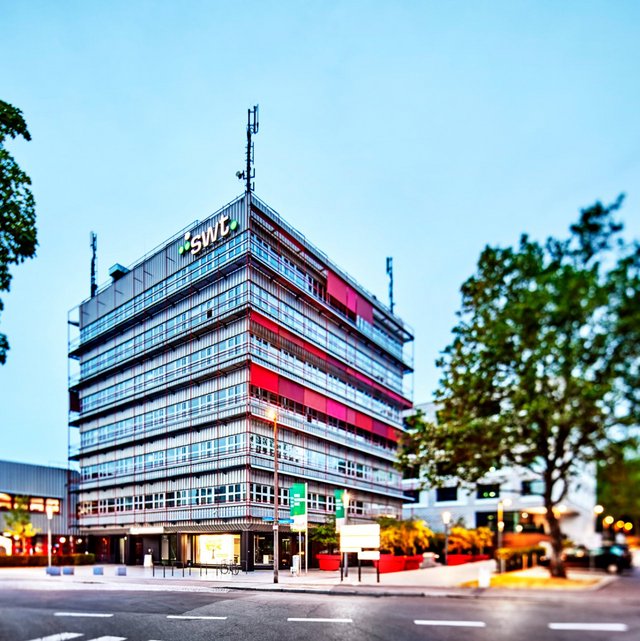 Ein moderner Bürokomplex mit einer Glasfassade in verschiedenen Farbtönen, darunter rot und grau. Das Gebäude hat mehrere Stockwerke und ist umgeben von Bäumen und Strassenlaternen. Im Vordergrund gibt es eine gepflasterte Fläche, auf der sich Fahrradständer befinden. Im Hintergrund sind weitere moderne Gebäude sichtbar, und die Umgebung scheint gut beleuchtet zu sein. Der Himmel ist klar und zeigt eine Dämmerungsstimmung.