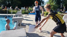 Zwei Männer in Sportkleidung stehen am Schwimmbecken, ein Mann sitzt im Wasser. Eine Person übergibt Unterlagen.
