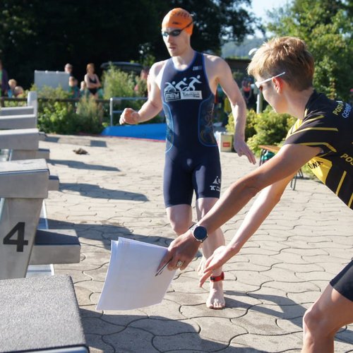 Zwei Männer in Sportkleidung stehen am Schwimmbecken, ein Mann sitzt im Wasser. Eine Person übergibt Unterlagen.