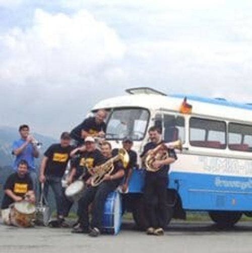 Eine Gruppe von sieben Musikern posiert vor einem blauen und weißen Bus in einer ländlichen Umgebung.