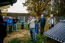 Eine Gruppe von Menschen steht im Freien, umgeben von Bäumen und einem Zaun. Im Vordergrund hält ein Mann mit Mikrofon in der Hand einen Vortrag und deutet mit der anderen Hand auf etwas. Die Zuhörer, darunter Männer und Frauen in unterschiedlichen Kleidungsstilen, hören aufmerksam zu. Einige Personen haben Notizblätter oder Tablets in der Hand. Im rechten Bildbereich sind Solarkollektoren zu sehen. Der Himmel ist bewölkt, und es ist herbstlich, mit einigen gelben Blättern an den Bäumen.