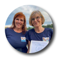 Zwei Frauen stehen nebeneinander und lächeln. Sie tragen dunkelblaue T-Shirts und haben Namensschilder auf der Brust. Im Hintergrund ist ein bewölkter Himmel zu sehen. Eine der Frauen hält ein Dokument in der Hand.