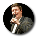Ein Mann mit braunen Haaren hält ein Mikrofon und lächelt, während er spricht. Er trägt einen dunkelgrauen Blazer mit einem karrierten Muster. Der Hintergrund ist schwarz, und das Bild hat eine runde Form.